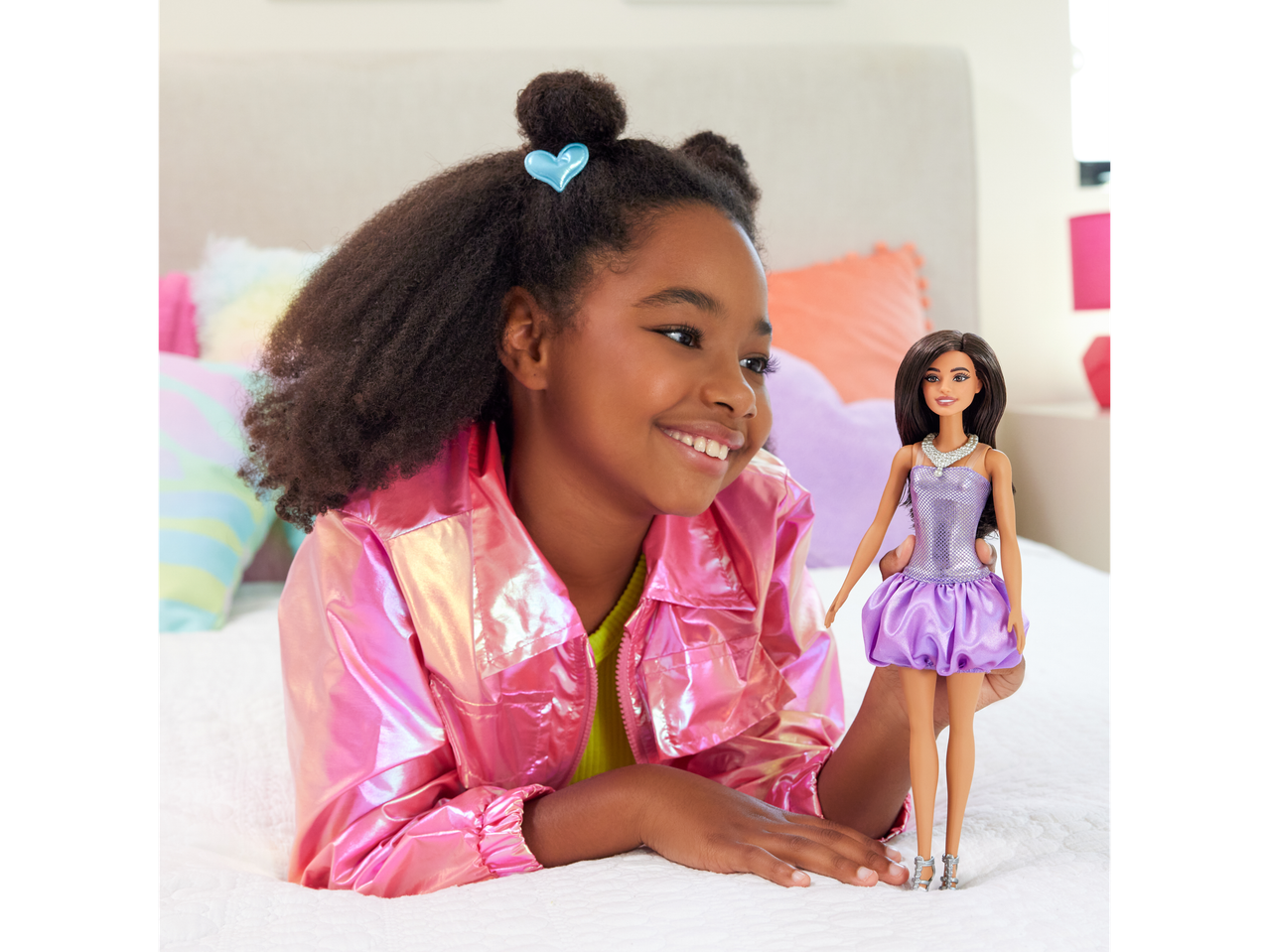 A girl in a pink jacket smiles at a Barbie doll in a purple dress.