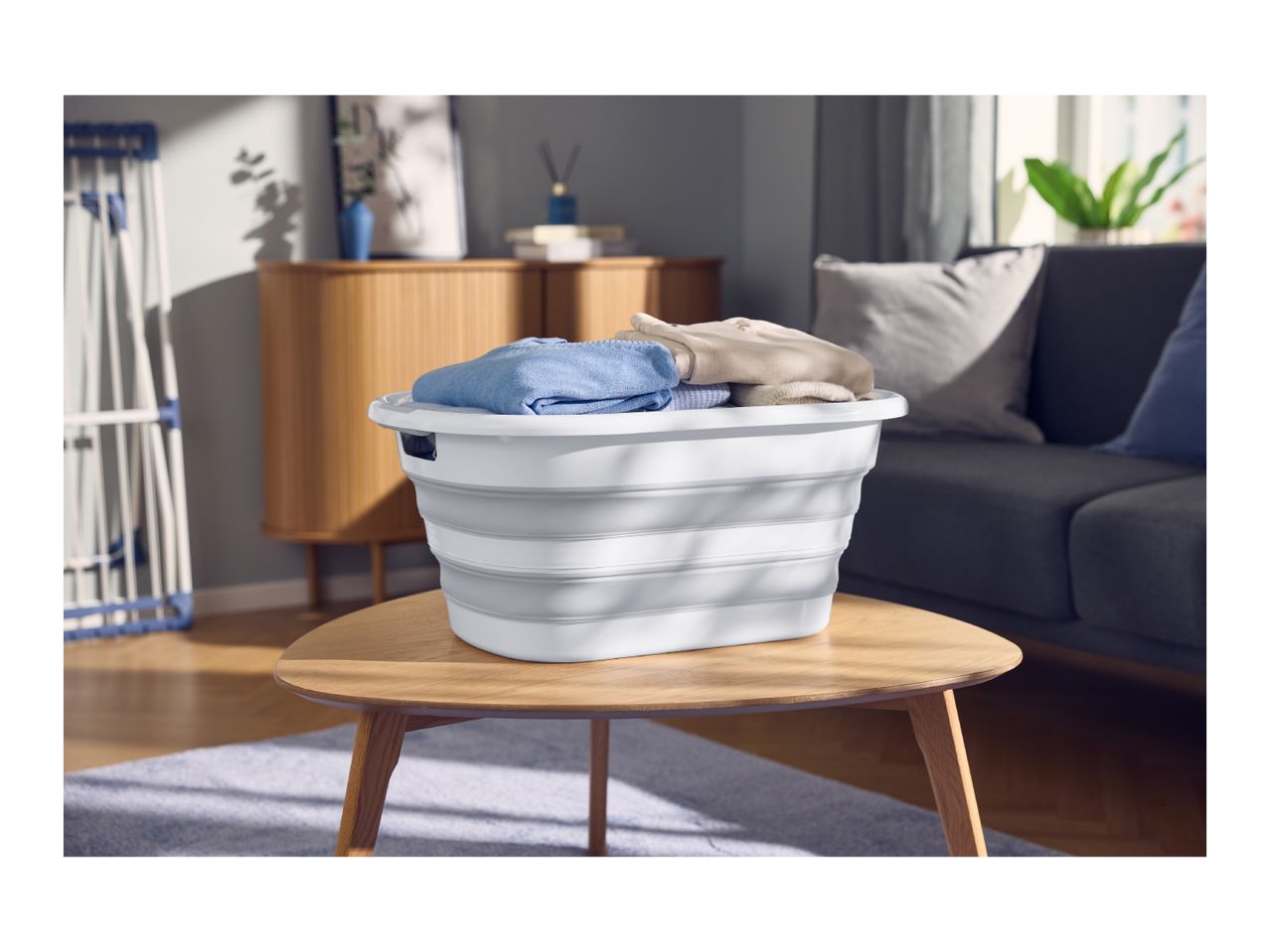 A white collapsible laundry basket filled with folded clothes on a wooden coffee table.