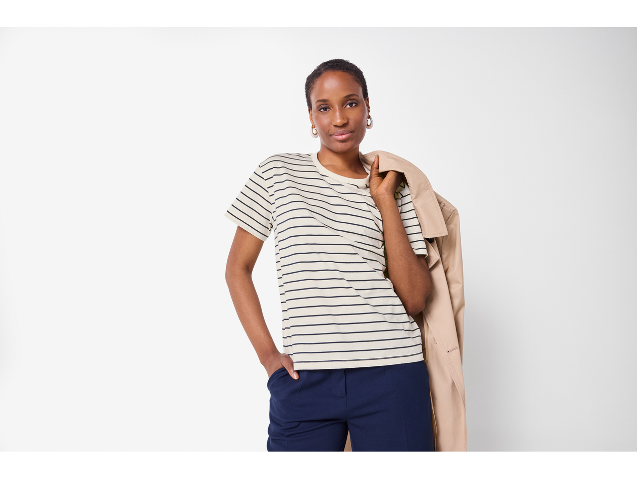 Mulher com t-shirt ESMARA às riscas, calças azuis e gabardine bege ao ombro.