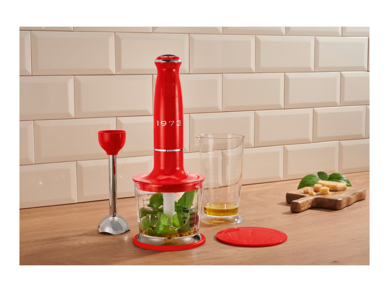 Red hand blender set with chopper, beaker, and whisk attachment on a wooden countertop