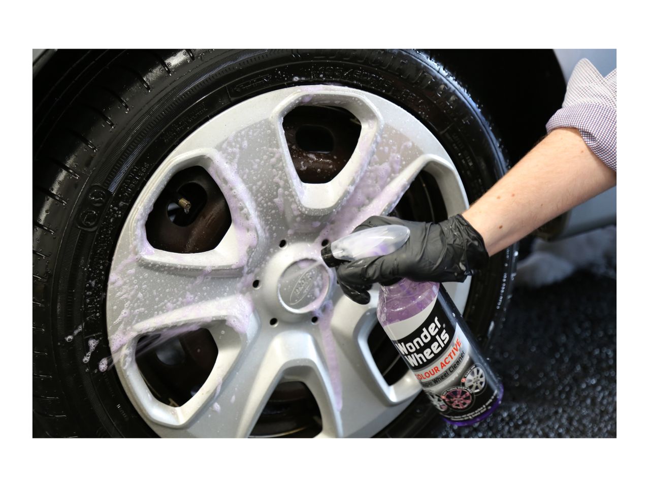 A person cleaning a car wheel with a spray bottle of wheel cleaner, creating purple foam.