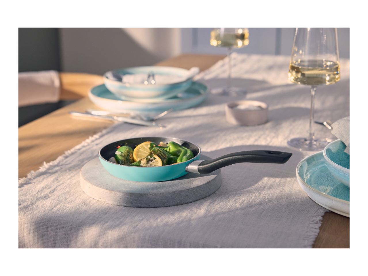 A small turquoise frying pan with green peppers and lemon slices on a table set for dining.