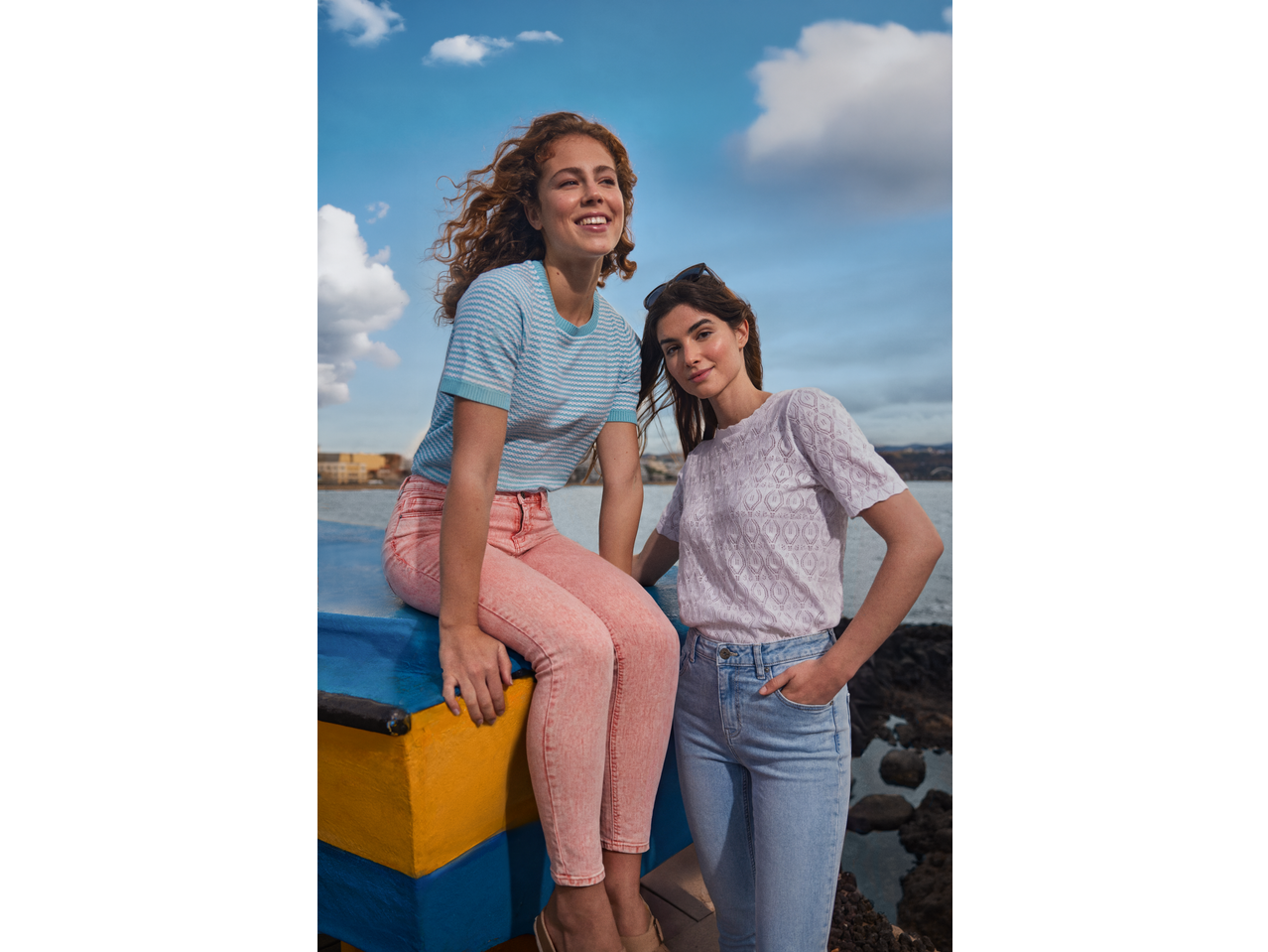 Duas mulheres vestindo blusas Esmara de malha aberta e jeans, sentadas perto da água.