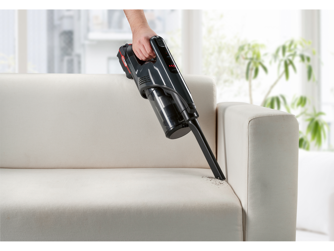 Ръка държи безжичен прахосмукачка Vorwerk Kobold VB100, докато чисти бял диван.