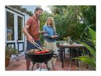 Man grilling meat and vegetables, woman holding a loaf of bread, outdoor dining.
