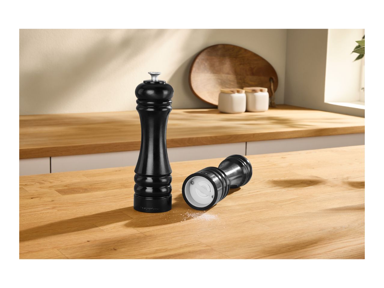 Two black pepper mills on a wooden kitchen counter, one standing, one lying down with salt.