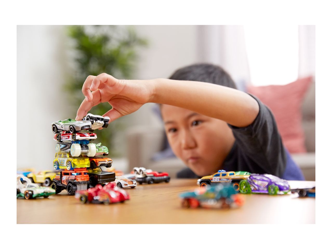 Child stacking toy cars on a wooden table, with more cars scattered around.
