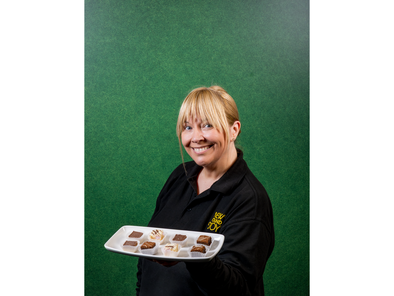 A woman in a black polo shirt holds a tray of brownies and other small desserts.