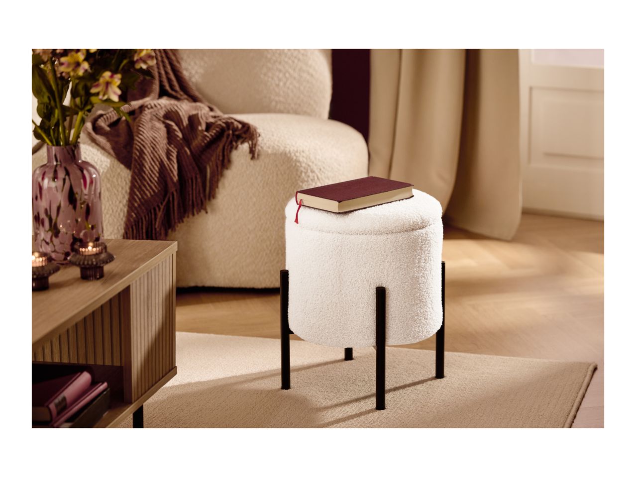 White bouclé ottoman with black legs, a red book on top, in a cozy living room.