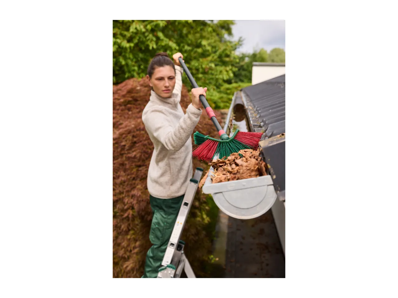 A woman on a ladder uses a telescopic gutter cleaner to clear leaves from a roof gutter.