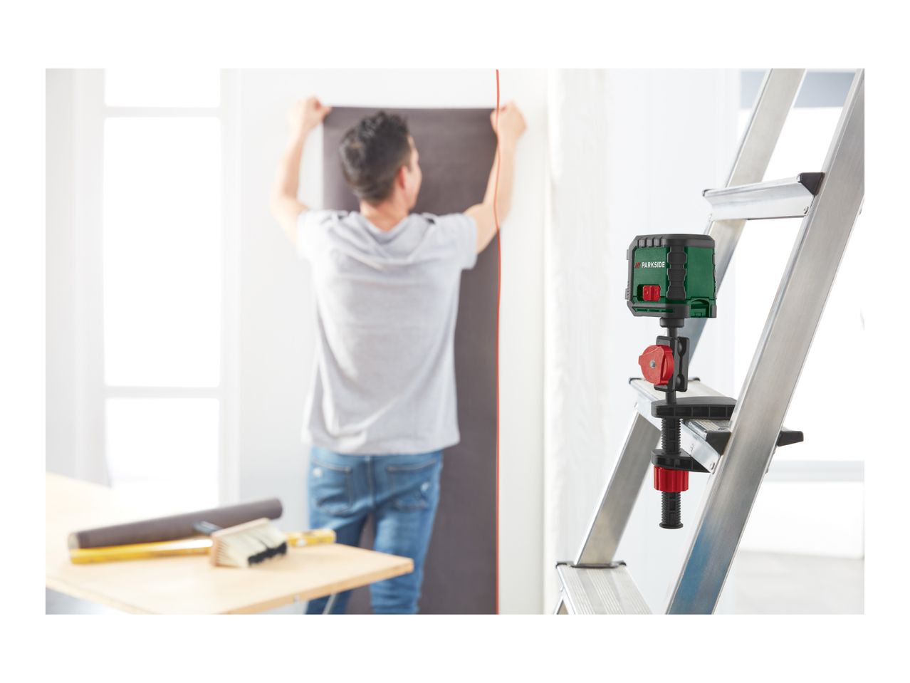 A laser level attached to a ladder, projecting a red line onto a wall where a person is hanging wallpaper.