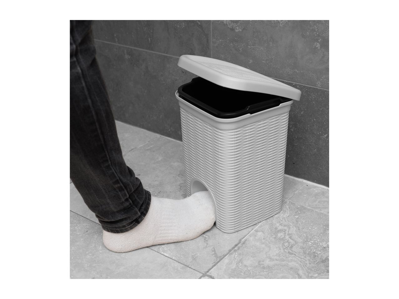 Foot opening a white pedal bin with a black inner bucket, against a grey tiled wall.