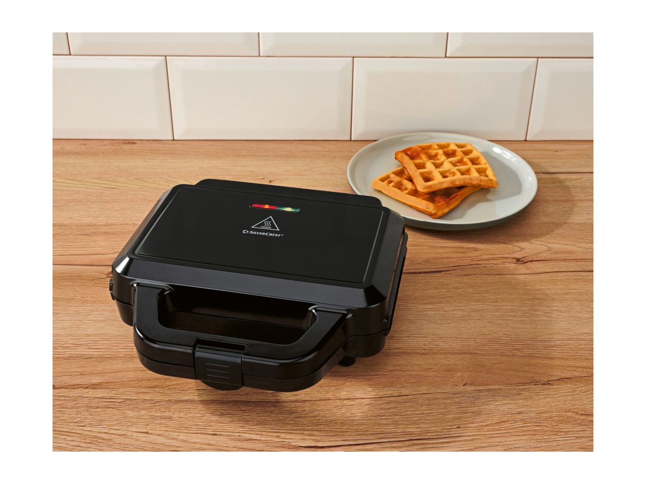 Black waffle maker on a wooden counter with two waffles on a plate in the background.
