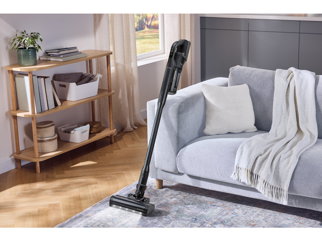 Kabelloser Staubsauger auf einem Teppich in einem Wohnzimmer mit Sofa und Holzregal.