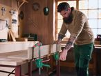 Man in a workshop using clamps to secure wood on a workbench, surrounded by tools.