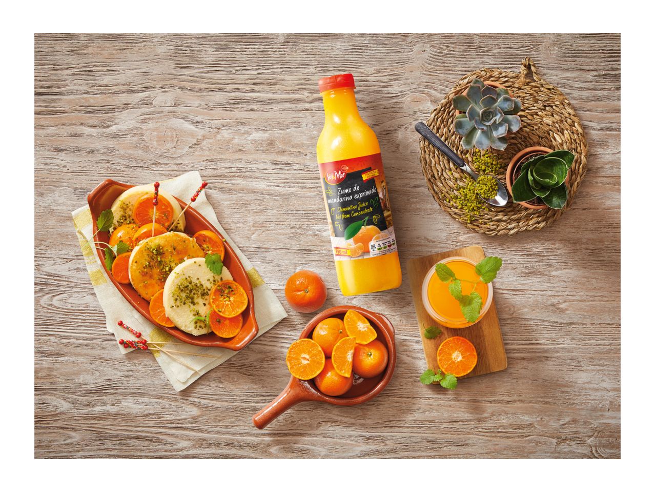 A bottle of clementine juice, a glass of juice, and a dish of clementines and dessert on a wooden table.