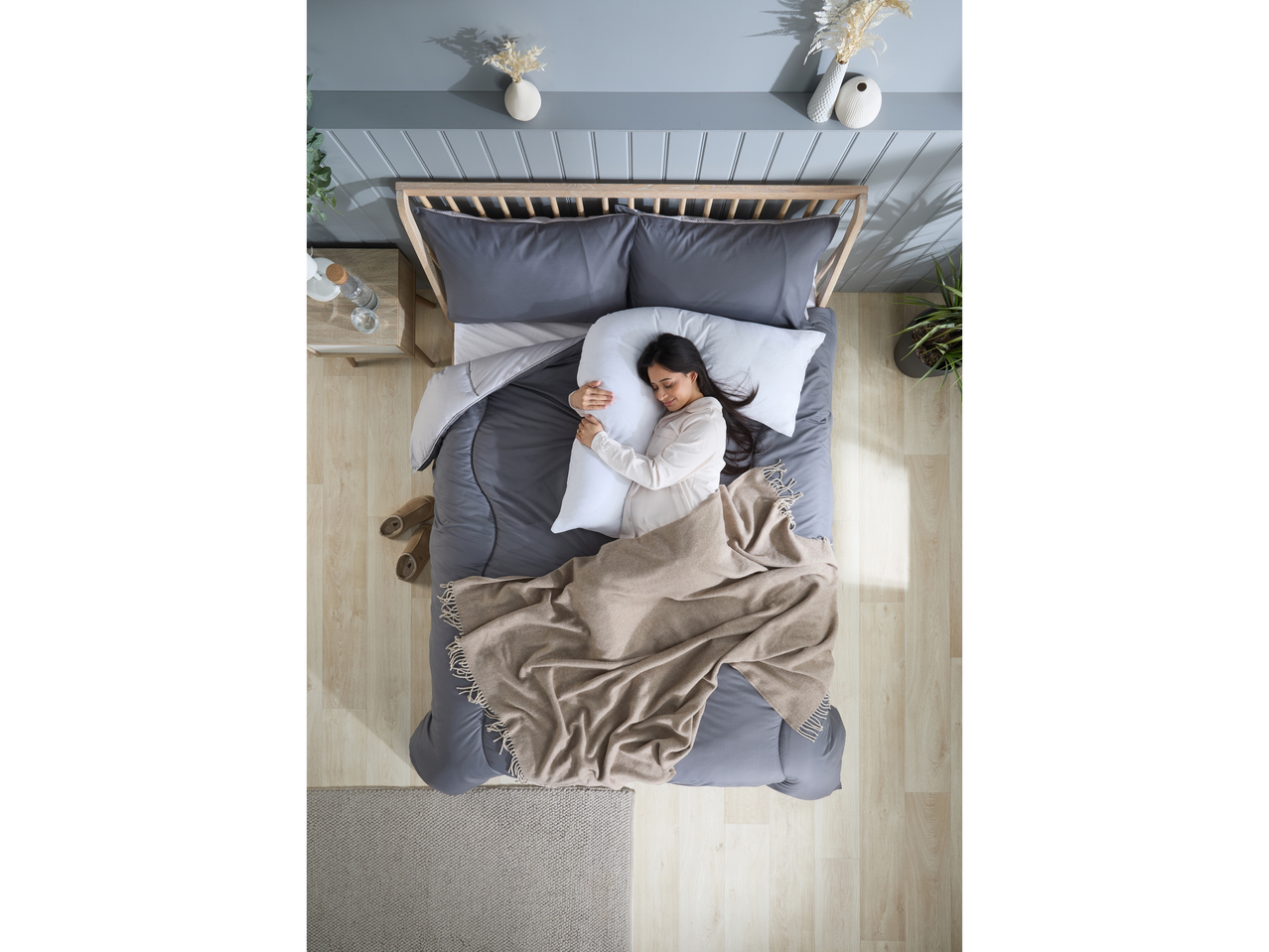 A woman sleeps soundly on a bed with a gray duvet and beige throw, using a body pillow.