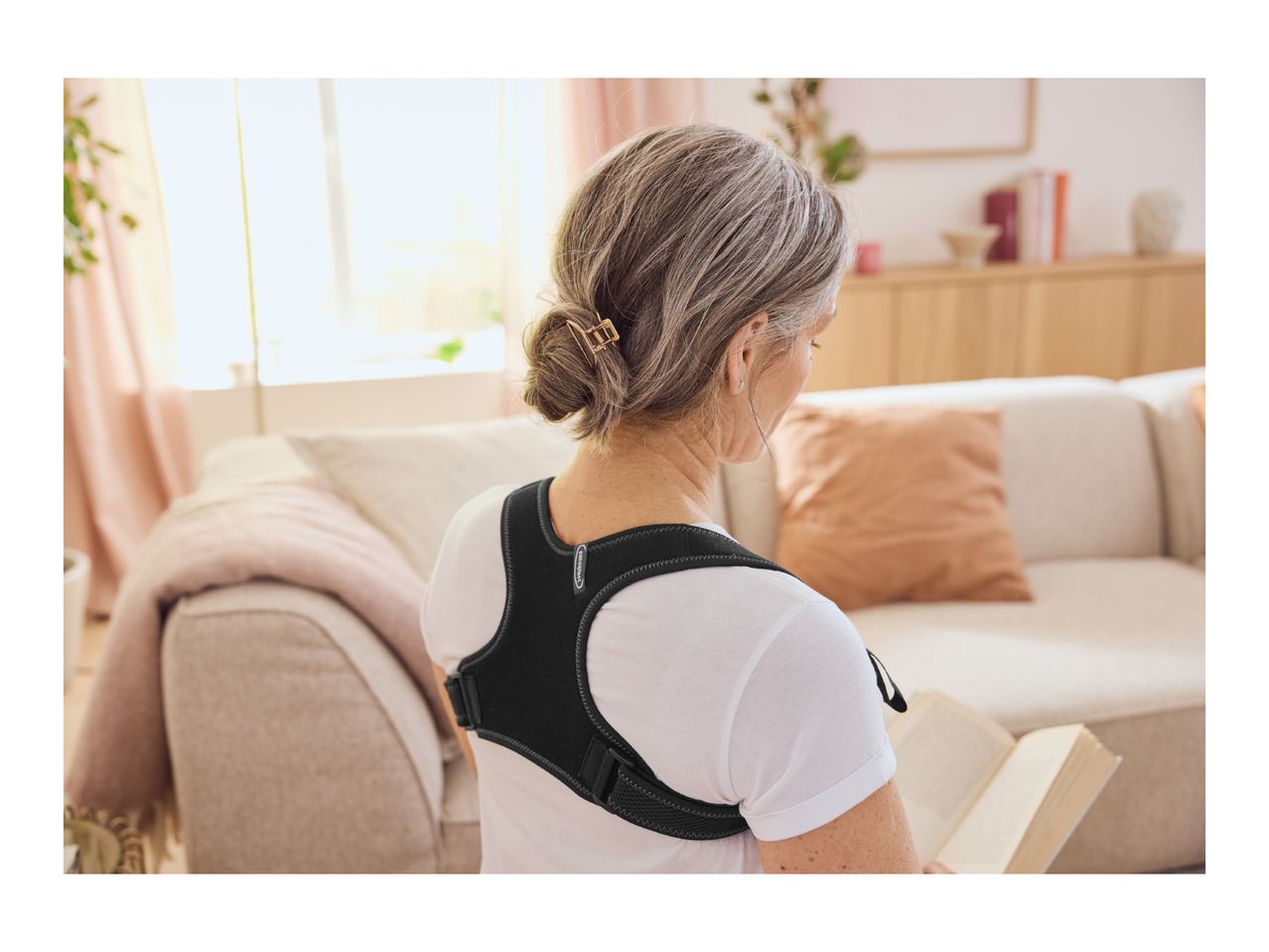 Woman wearing a posture corrector while reading a book on a sofa.