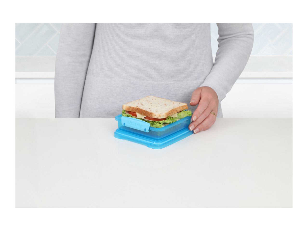 Person holding a blue sandwich container with a sandwich inside on a white table.