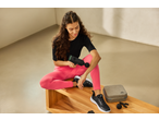 Mulher de leggings cor-de-rosa e camisola preta a massajar o braço com pistola de massagem, sapatilhas ao lado.