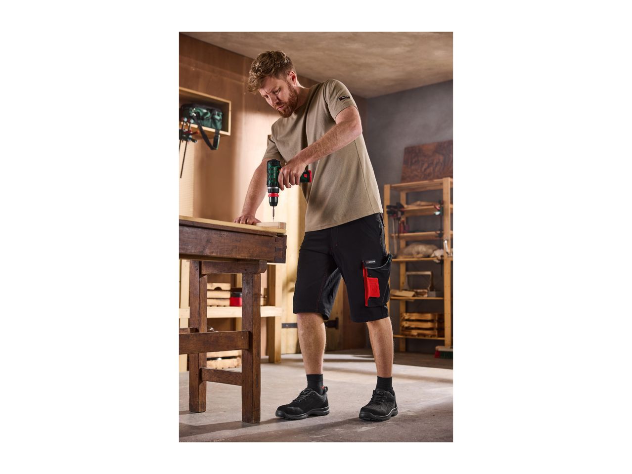 Man in work shorts and t-shirt drilling wood on a workbench in a workshop.