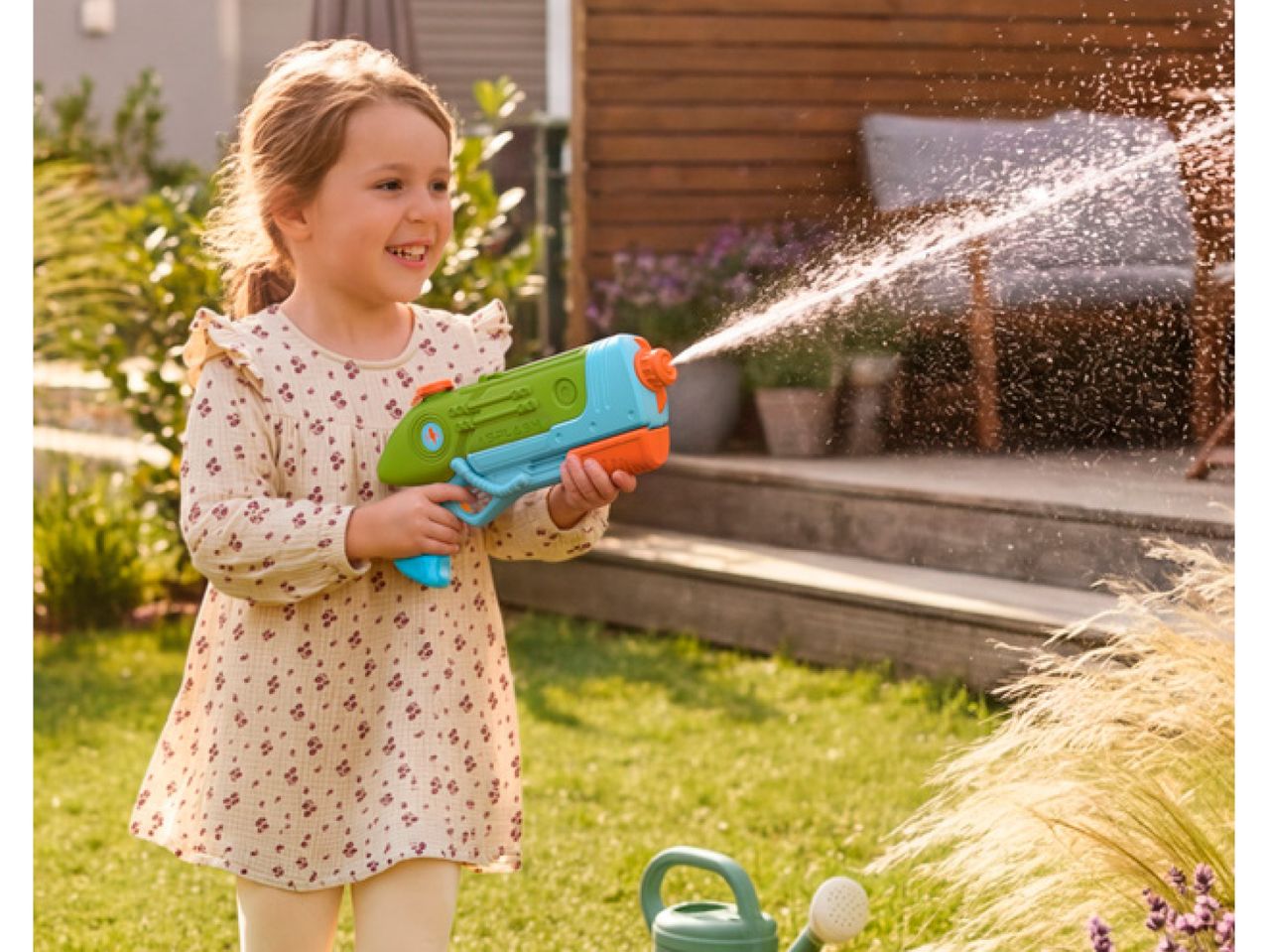 Smiling girl playing with a water blaster in a sunny garden.