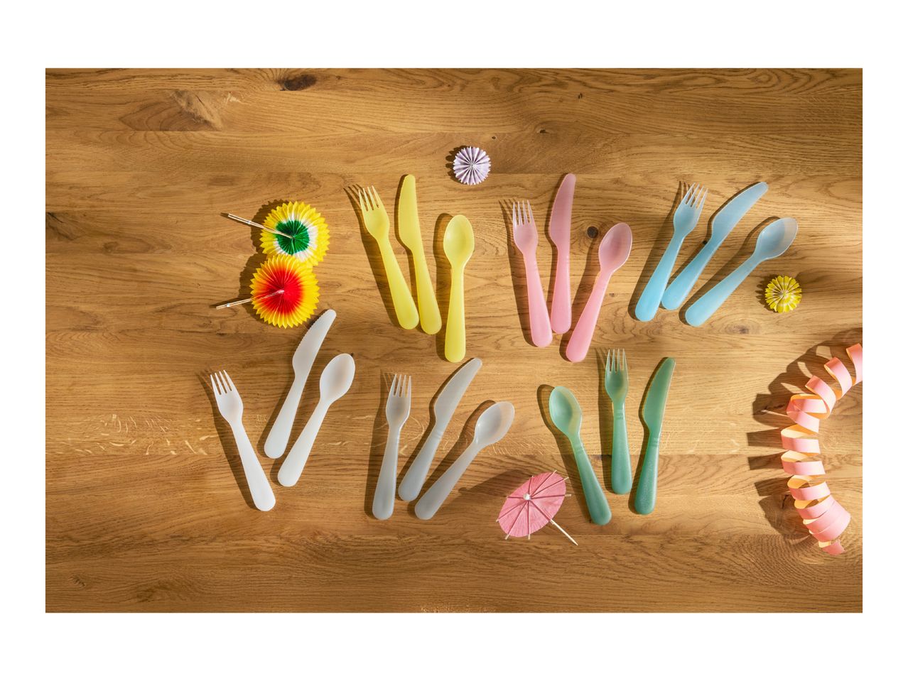 Colorful plastic cutlery sets in yellow, pink, blue, white, and green, with party decorations on a wooden table.