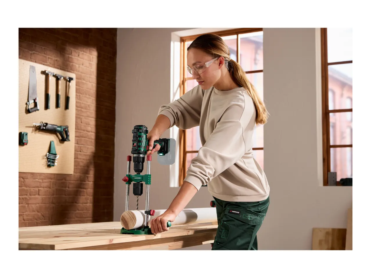 A woman using a drill guide attachment to drill a hole into a wooden post on a workbench.