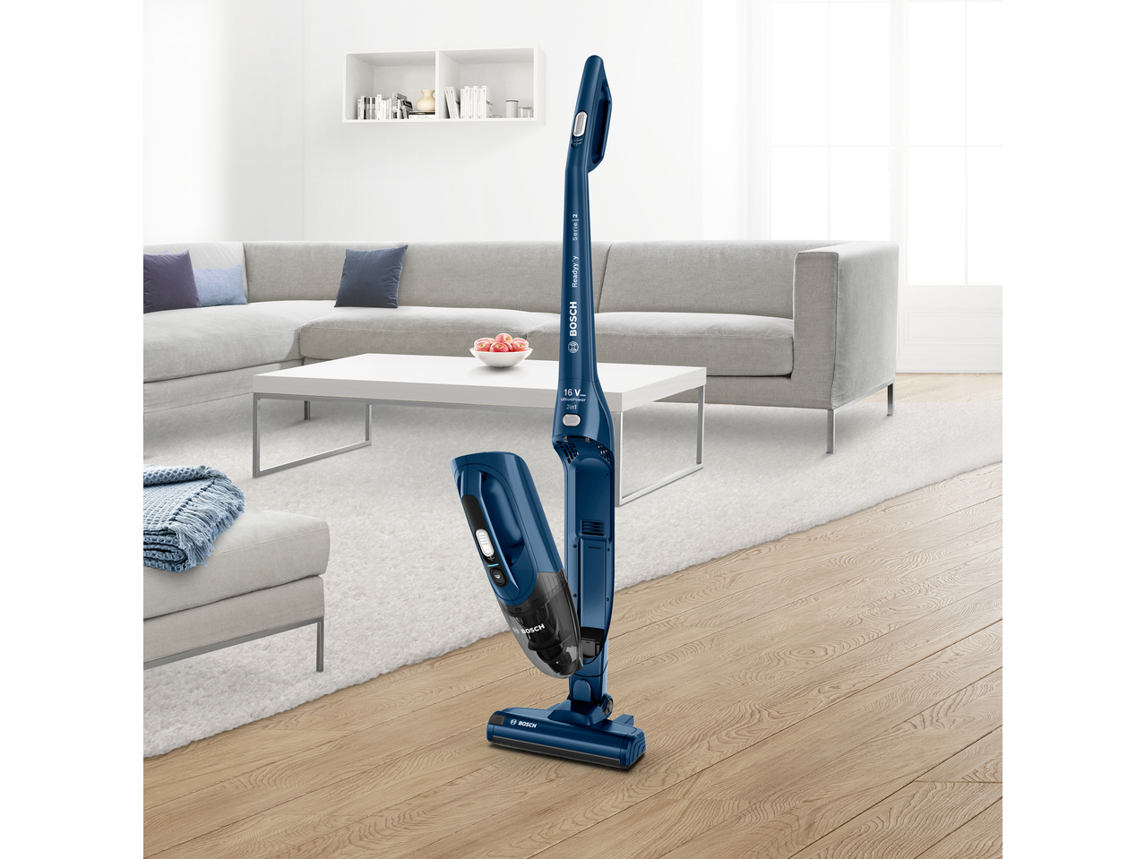 Blue cordless stick vacuum cleaner in a modern living room with a grey sofa and white coffee table.
