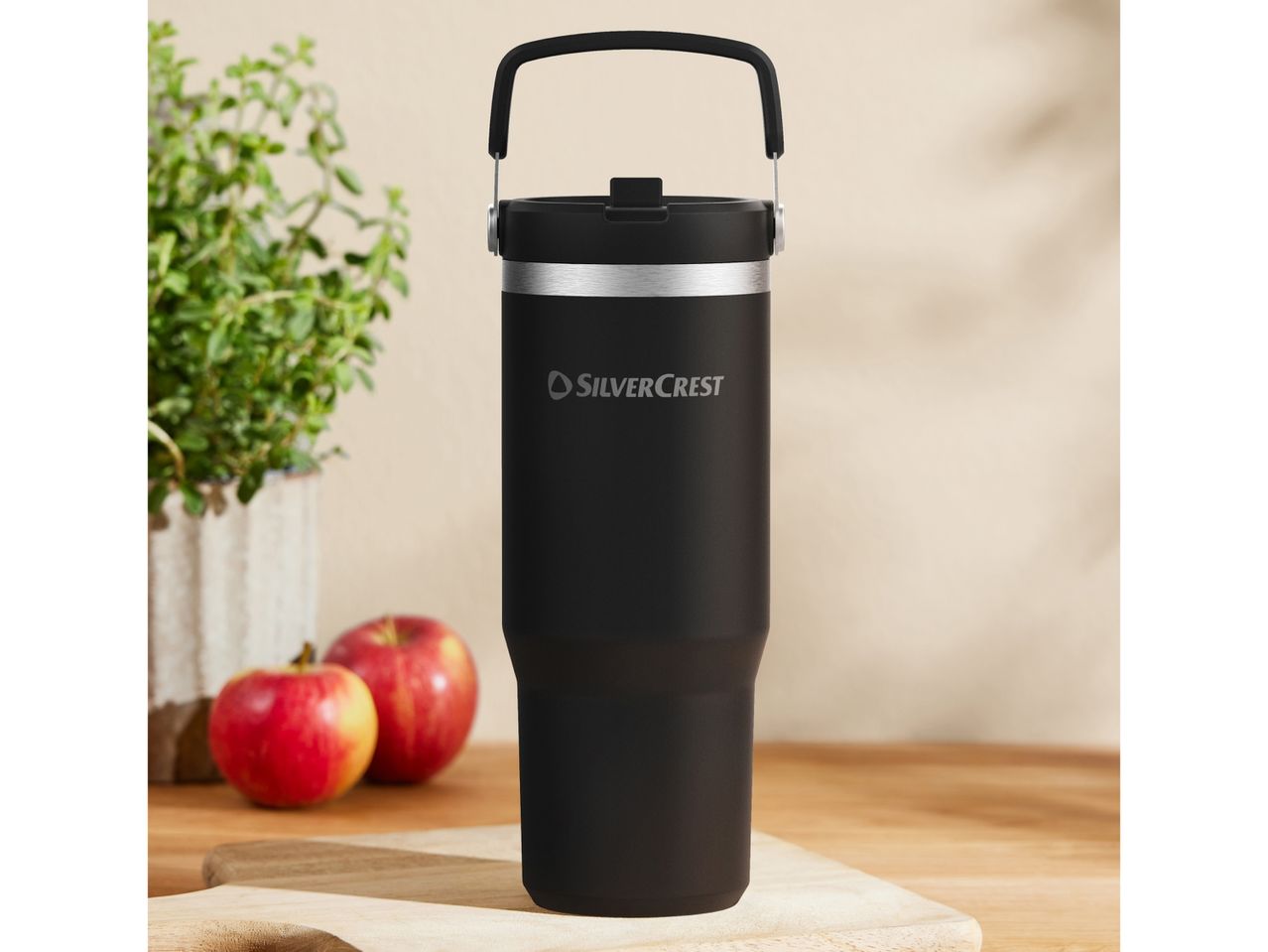 Black SilverCrest travel mug with handle, apples, and a plant on a wooden surface.