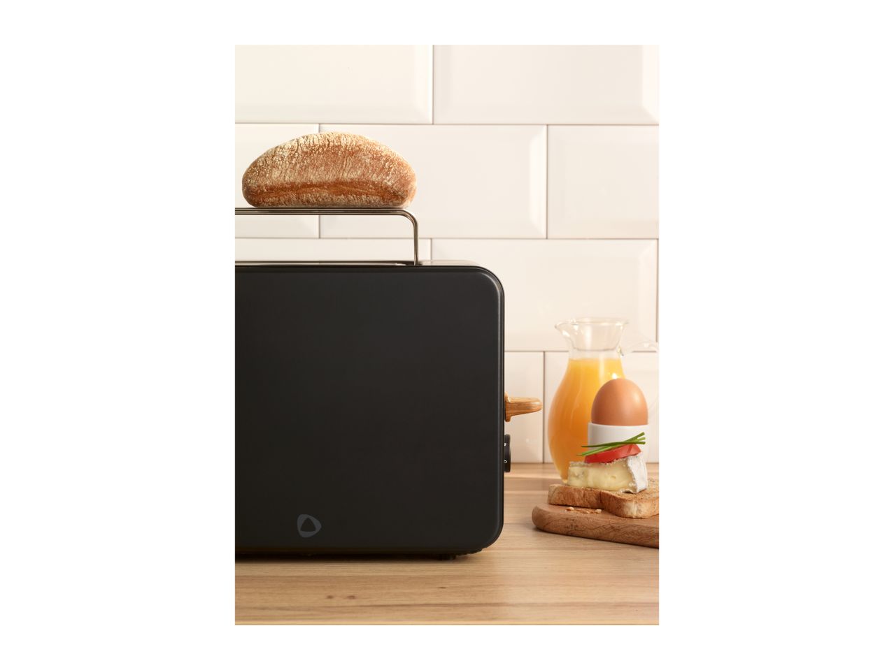 Black toaster with a bread roll on top, next to an egg, cheese, and orange juice.