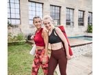 Two women in workout clothes carrying yoga mats, smiling and looking at each other.