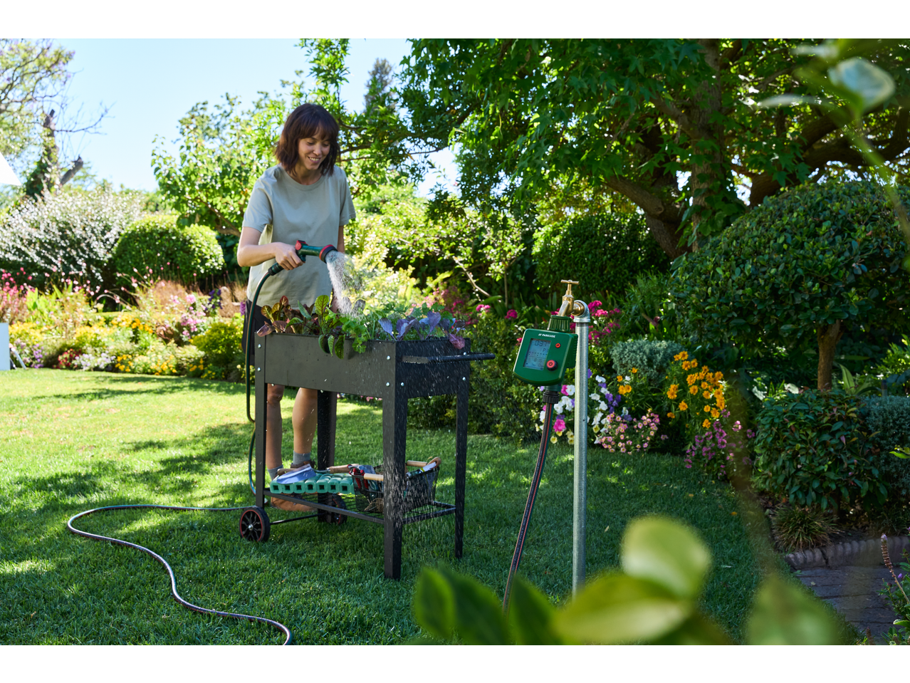 Femeie udând o jardinieră înălțată cu un temporizator de apă într-o grădină însorită.