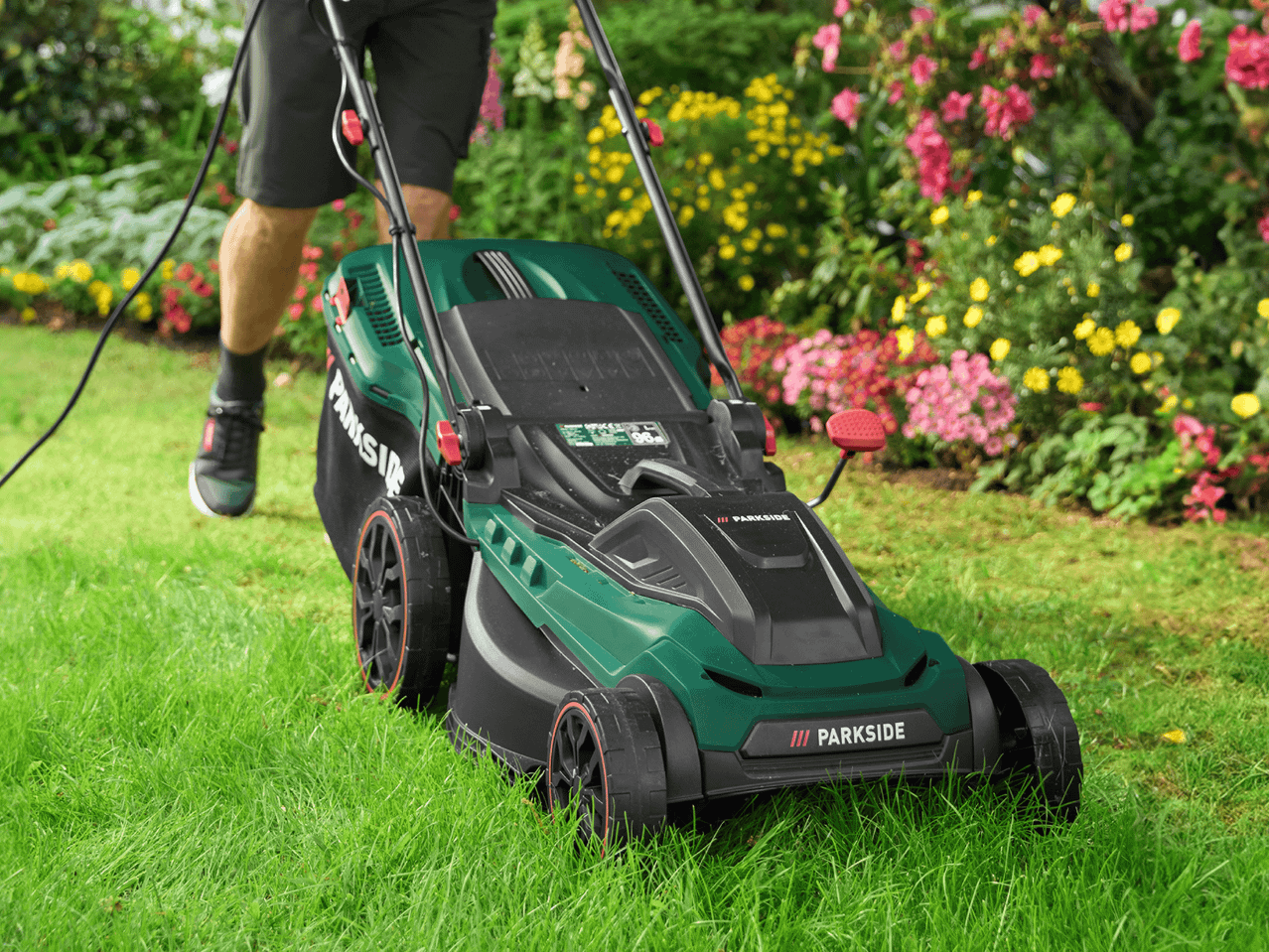 Eine Person mäht den Rasen mit einem grünen Rasenmäher vor einem blühenden Blumenbeet.