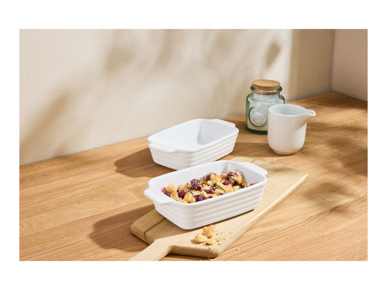Two white rectangular baking dishes, one filled with fruit crumble, on a wooden surface.