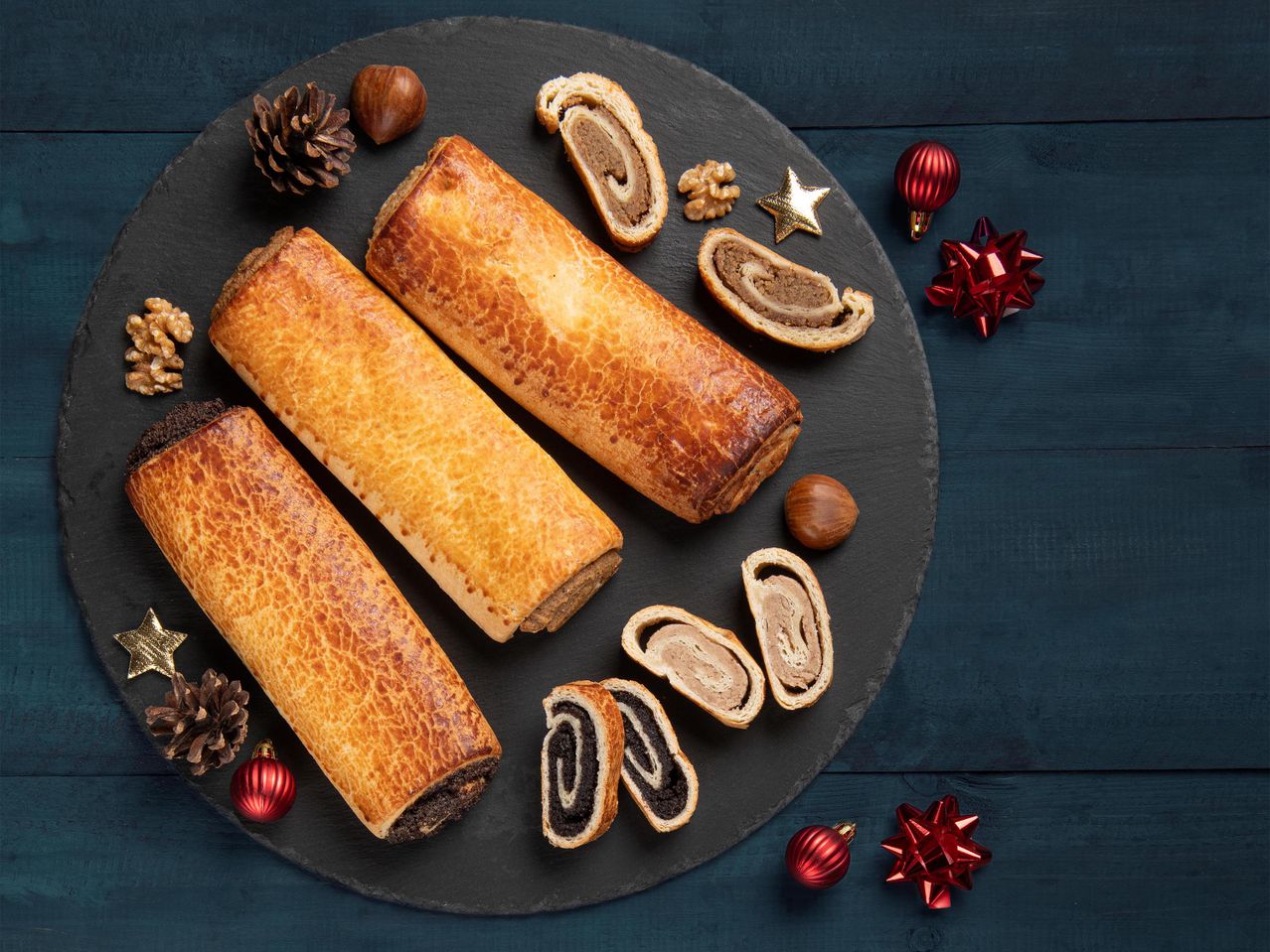 Három hagyományos magyar bejgli tekercs, kettő diós és egy mákos töltelékkel, sötét palatáblán karácsonyi díszekkel.