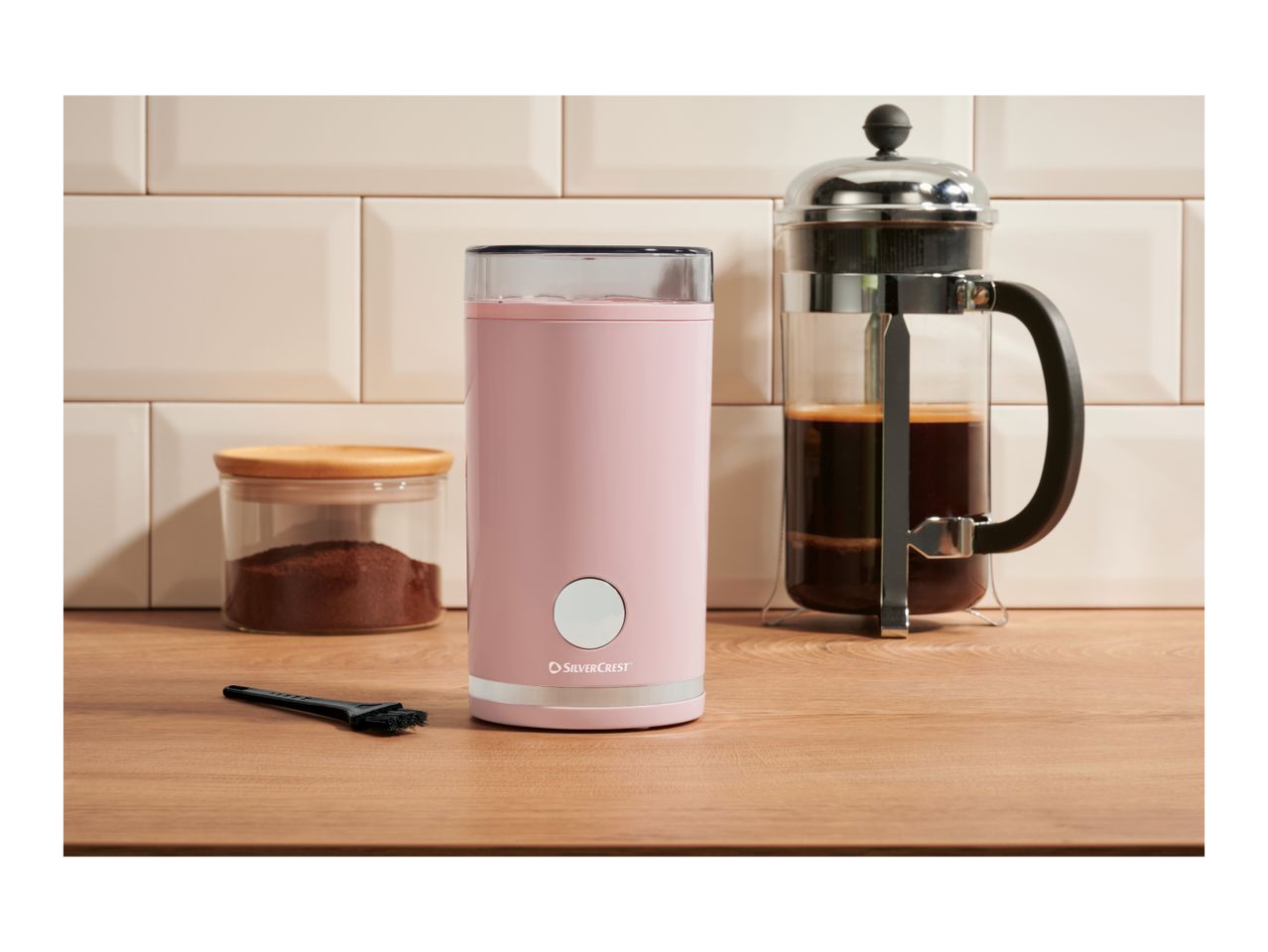 Pink coffee grinder, French press, and a jar of ground coffee on a wooden counter.