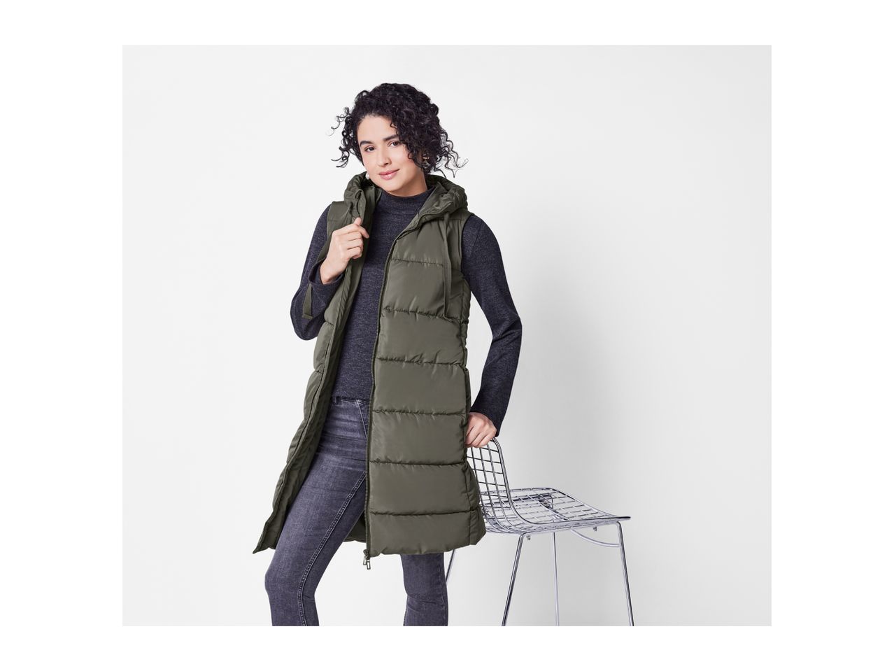 Woman in a long olive green puffer vest, dark gray long-sleeve top, and dark jeans.