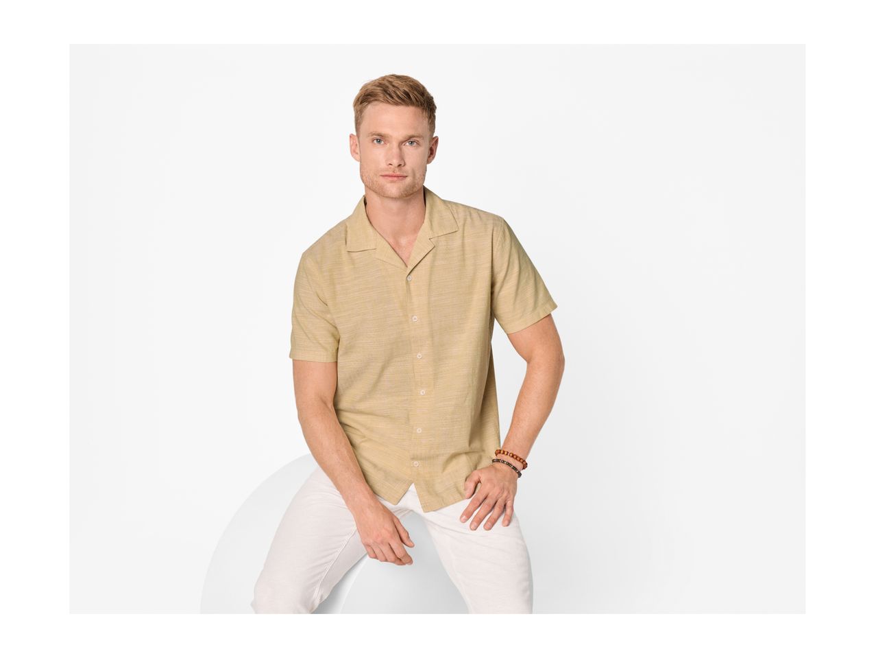 Man wearing a short-sleeved beige shirt and white trousers, sitting on a white surface.