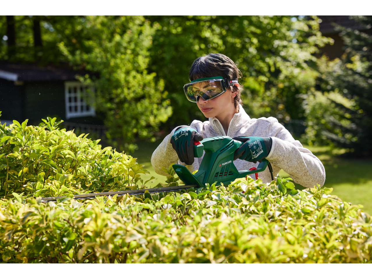 A woman uses a Parkside electric hedge trimmer to trim a hedge in her garden.