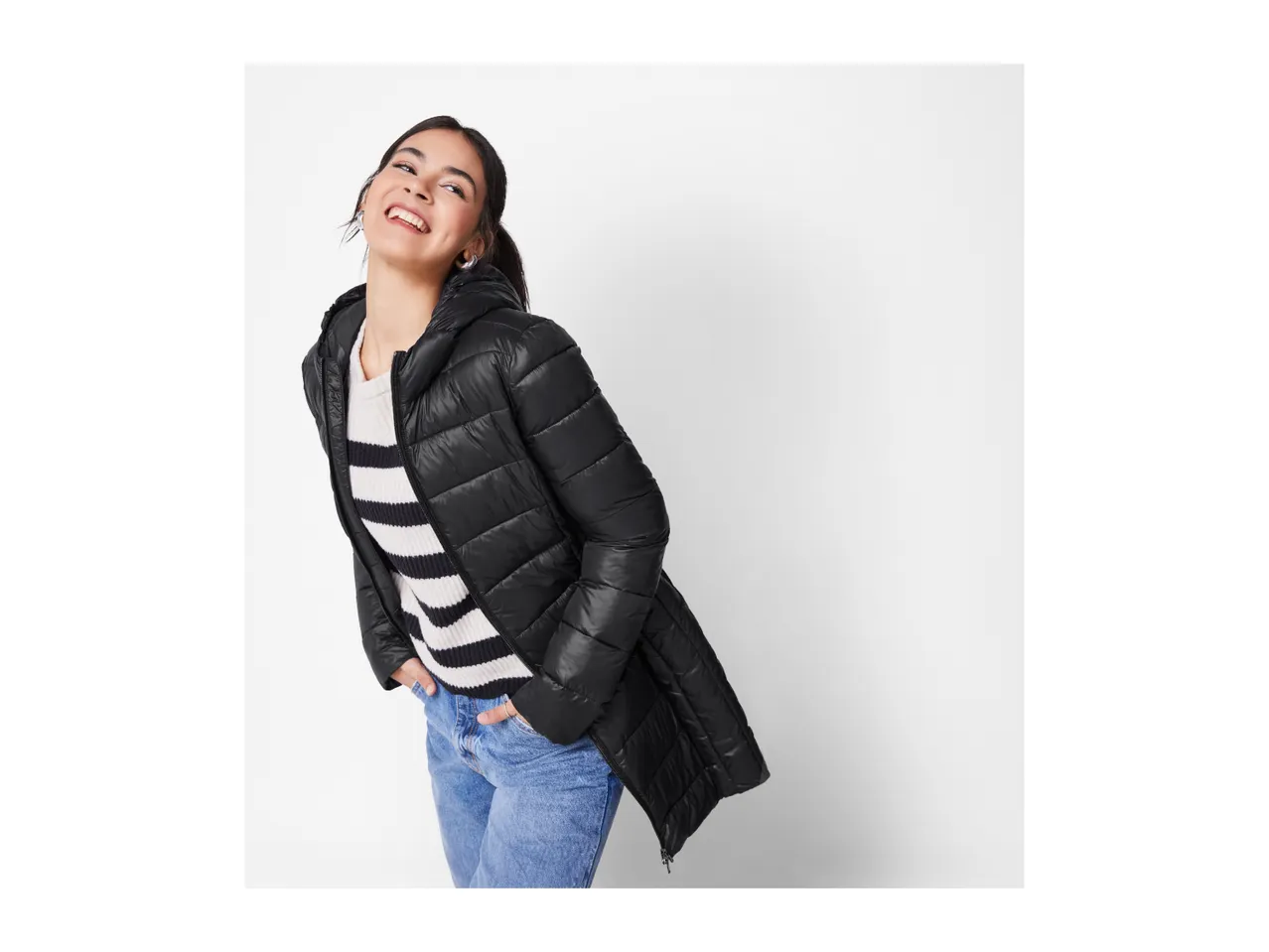 A woman in a black puffer jacket, striped sweater, and blue jeans smiles.