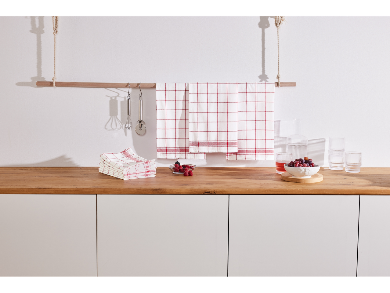 Serviettes de cuisine à carreaux rouges, ustensiles et verres sur un comptoir en bois.