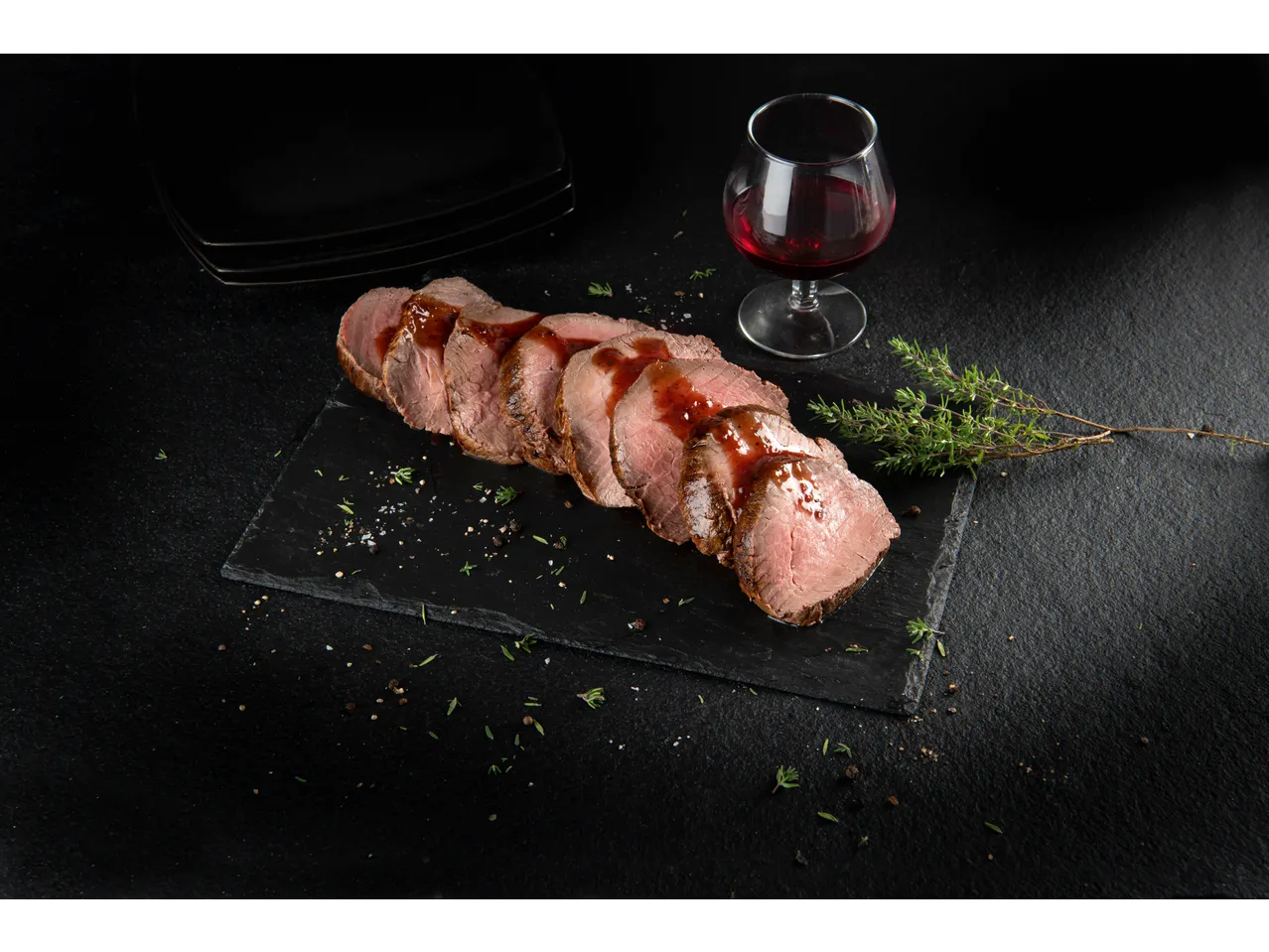 Sliced roast beef with sauce, herbs, and a glass of red wine on a dark background.