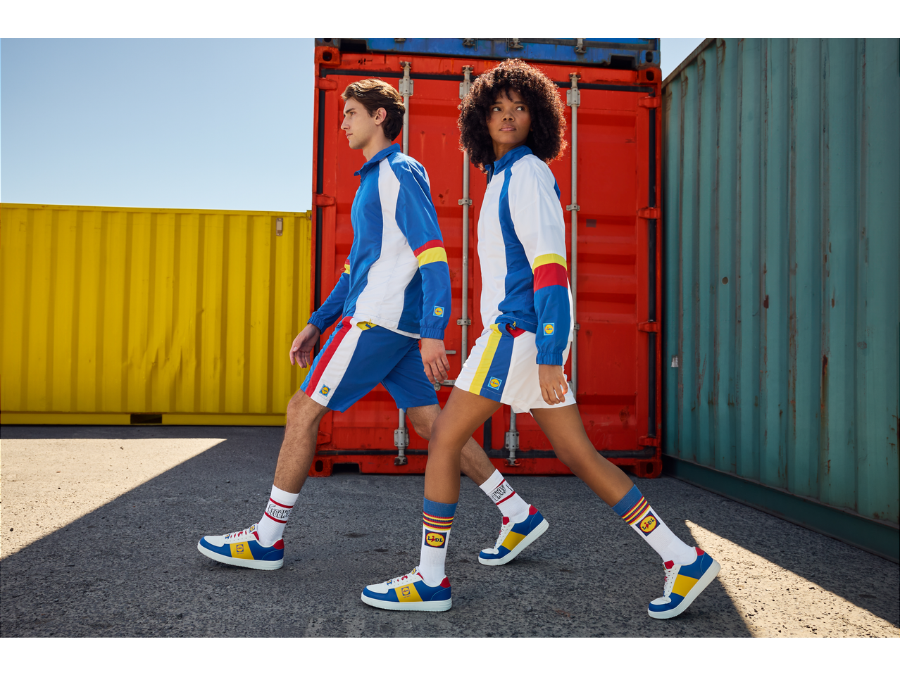 Two models in tracksuits, shorts, socks, and sneakers, walking past colorful shipping containers.