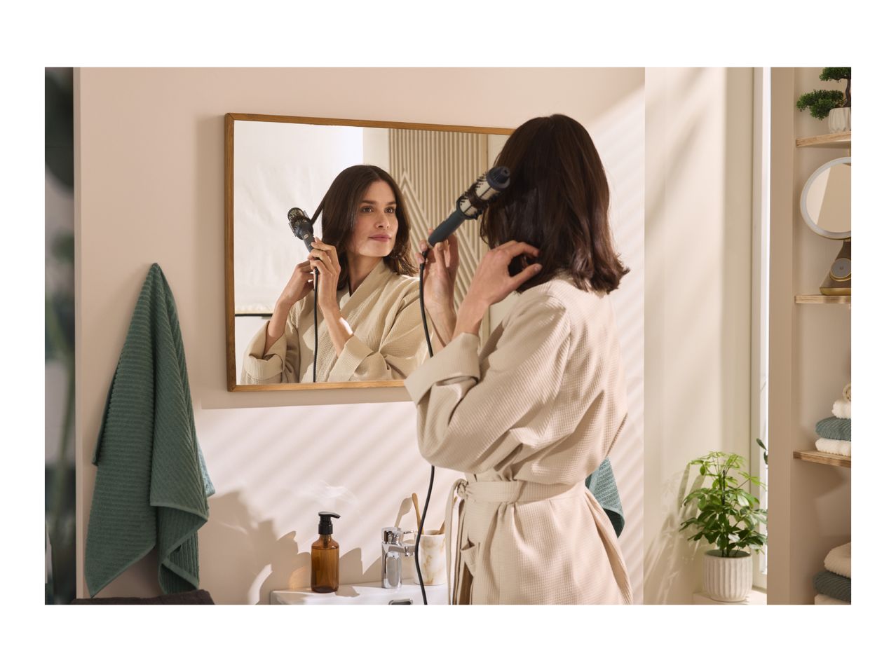 Woman in bathrobe styling her hair with a hot brush in a bathroom.