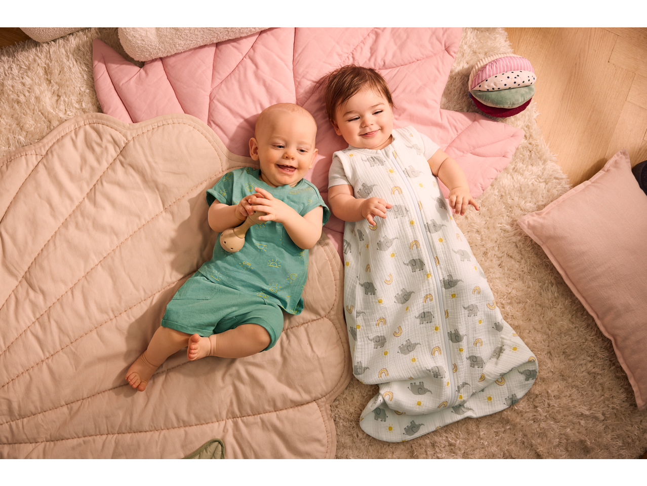 Doi bebeluși fericiți stau pe pături, unul într-un body LUPILU verde și celălalt într-un sac de dormit LUPILU.