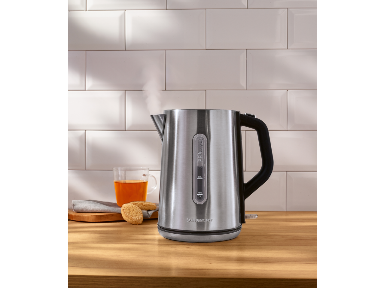 Steaming electric kettle on a wooden counter with a cup of tea and cookies.