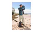 Man in outdoor jacket and cargo pants looking through binoculars on a sandy beach.