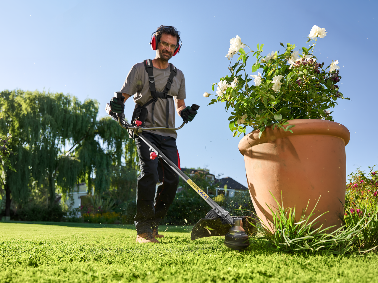 Άνδρας χρησιμοποιεί χορτοκοπτικό σε κήπο, φορώντας γυαλιά και ωτοασπίδες.