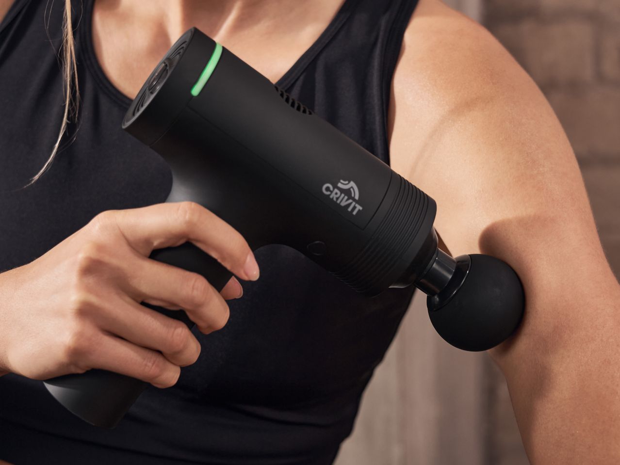 A person in a black sports top uses a black massage gun on their arm.